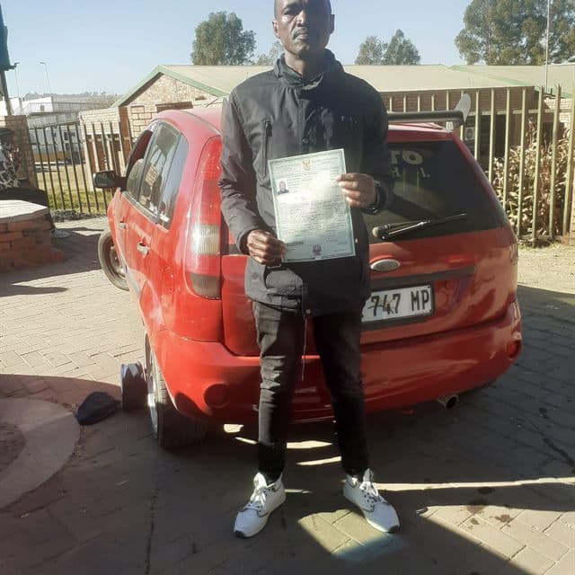 Phindile Z standing with a Ditheto Driving Academy training car and licence certificate.
